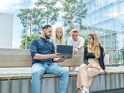 Auf dem Foto sind vier Studierende auf dem Bildungscampus zu sehen, die gemeinsam auf einen Laptop schauen.