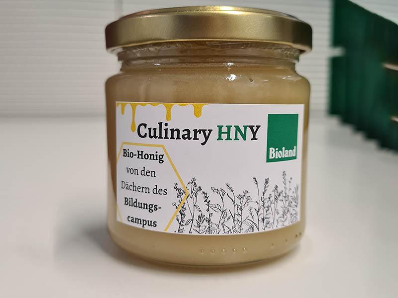 Das Foto zeigt ein Honigglas von Culinary HNY mit der Aufschrift "Bio-Honig von den Dächern des Bildungscampus".