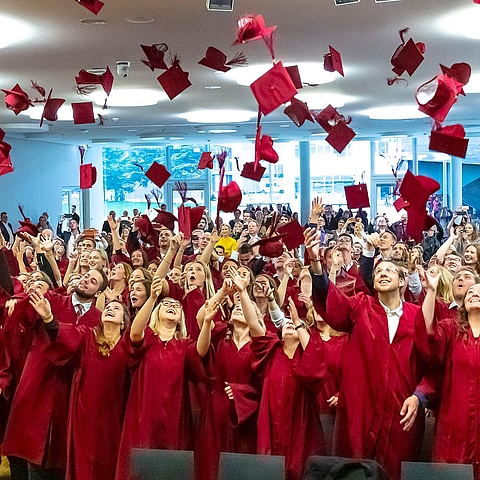 Auf dem Foto sind Absolventen und Absolventinnen der DHBW Heilbronn während ihrer Graduierungsfeier zu sehen. Sie tragen alle einen roten Talar und werfen rote Doktorhüte in die Luft.