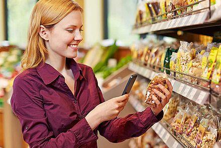 Auf dem Foto ist eine Frau zu sehen, die vor einem Einkaufsregal steht und ein Produkt mit dem Handy scannt.