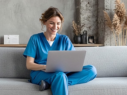Das Bild zeigt eine junge Frau, die mit einem Laptop in blauer Arbeitskleidung auf einem Sofa sitzt