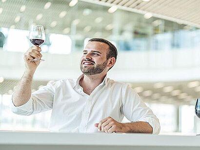 Auf dem Bild ist ein Mann mit einem Weinglas zu sehen. Er hält das Glas hoch und betrachtet es. 