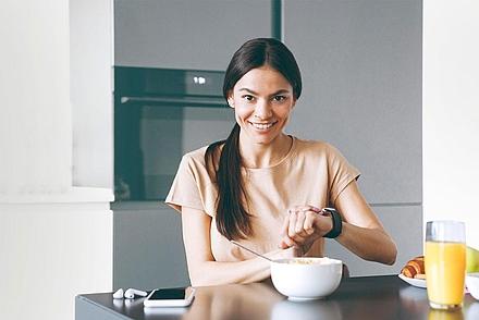 Eine junge Frau sitzt bei einem gesunden Frühstück am Tisch und fasst an ihre Smartwatch