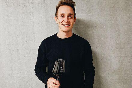 Der Absolvent des Studiengangs Wein-Technologie-Management (WTM) an der DHBW Heilbronn Nico Wiedemann steht vor einer grauen Wand und hält ein Weinglas halb gefüllt mit Rotwein in der Hand. Er trägt einen schwarzen Pullover. 
