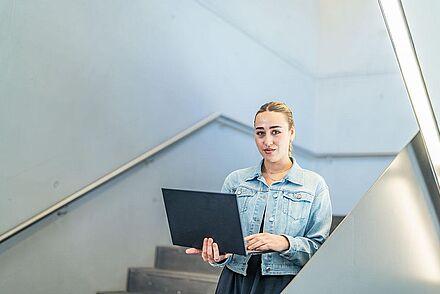 Auf dem Foto ist eine Frau zu sehen, die auf einer Treppe steht und einen Laptop in der Hand hält.