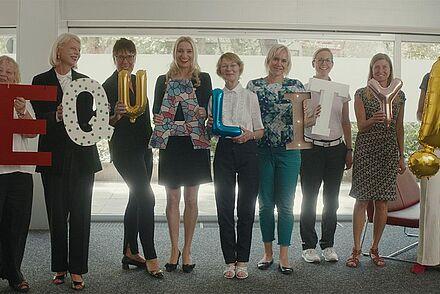 Auf dem Foto sind neun Frauen zu sehen, die nebeneinander stehen. Sie halten jeweils einen Buchstaben in der Hand und bilden das Wort EQUALITY!