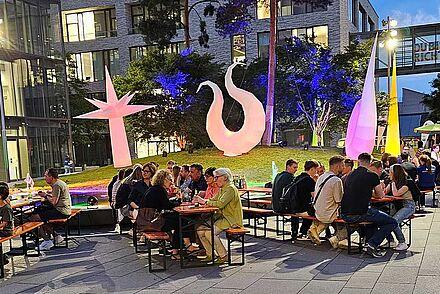 Hier sitzen viele Besucher*innen an Biertischen vor der Bühne auf dem Bildungscampus Mitte und hören der Band zu. Der Bildungscampus ist bunt illuminiert.
