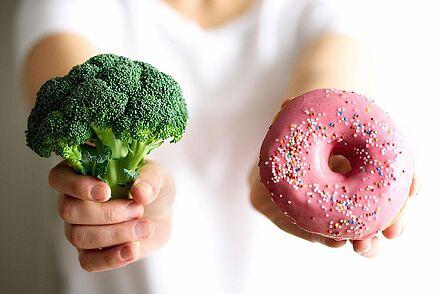 Auf dem Foto ist eine Frau zu sehen, die einen Brokkoli und einen Doughnut in die Kamera hält. Das soll die Auswahl der verschiedenen Lebensmittel für die ganz persönliche Ernährung symbolisieren.