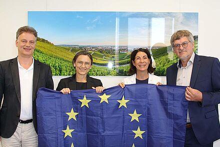 Auf dem Bild stehen vier Personen nebeneinander und halten dabei eine Europaflagge. Von links nach rechts: Prof. Dr. Thomás Bayón, Prof. Dr. Martina Klärle, Prof. Dr. Nicole Graf und Raimund Hudak.