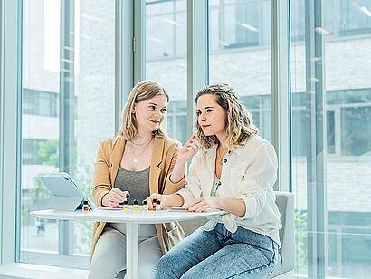 Das Foto zeigt zwei Frauen die an einem Tisch sitzen und kleine Glasfläschchen in der Hand halten. Auf dem Tisch stehen noch sechs weitere Glasfläschchen. Es sieht so aus, als würden sie einen Geruchstest durchführen.