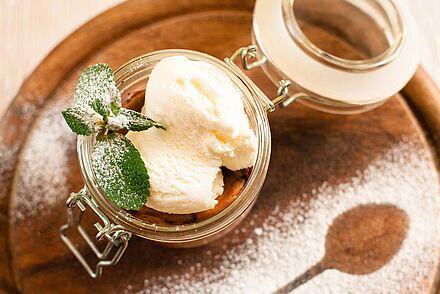 Zwei Kugeln Eis in einem Weckglas mit zwei Minzblättern garniert und mit Puderzucker bestäubt. Neben dem Weck-Glas ist der Abdruck eines Löffels zu sehen. 