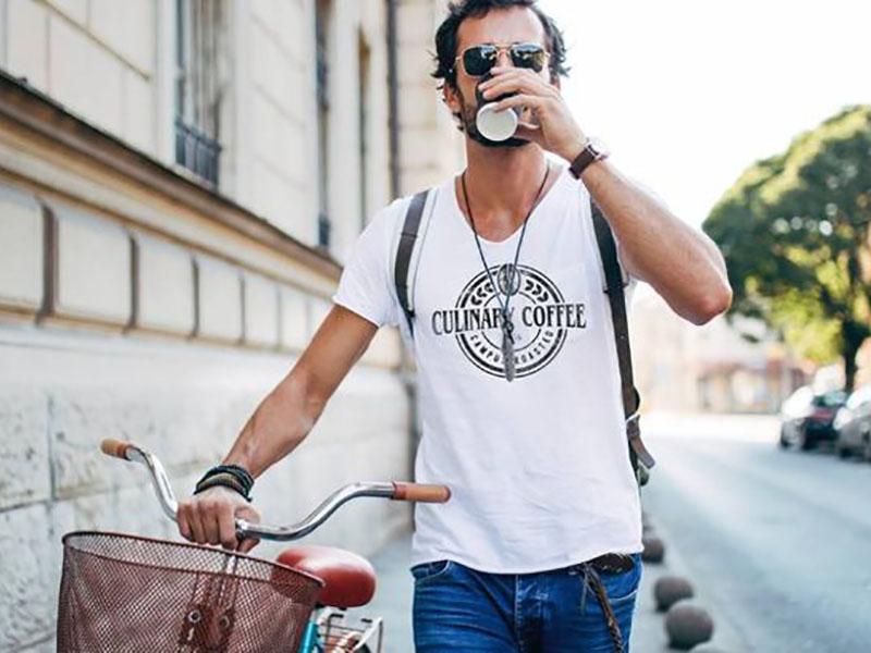 Auf dem Foto ist ein Mann zu sehen, der draußen auf dem Gehweg läuft, sein Fahrrad schiebt und einen Kaffee trinkt. Er trägt ein weißes T-Shirt, auf dem das Logo von Culinary Coffee aufgedruckt ist.