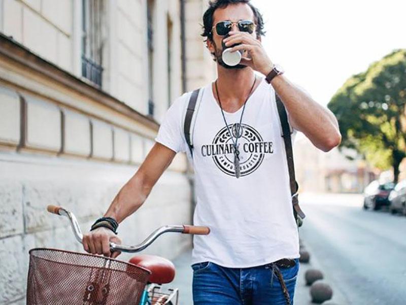 Culinary Coffee T-Shirt Auf dem Foto ist ein Mann zu sehen, der draußen auf dem Gehweg läuft, sein Fahrrad schiebt und einen Kaffee trinkt. Er trägt ein weißes T-Shirt, auf dem das Logo von Culinary Coffee aufgedruckt ist.