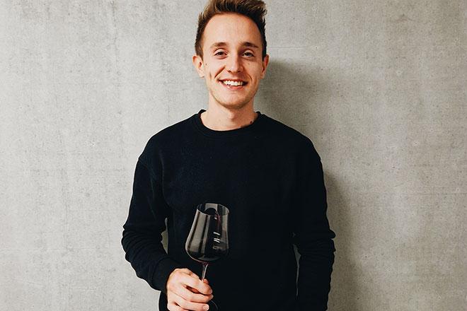 Der Absolvent des Studiengangs Wein - Technologie - Management an der DHBW Heilbronn Nico Wiedemann hält ein Glas Rotwein in der Hand, die andere Hand ist in der Hosentasche und er lächelt nonchalant in die Kamera. Er trägt einen schwarzen Pullover und steht vor einer grauen Wand. 