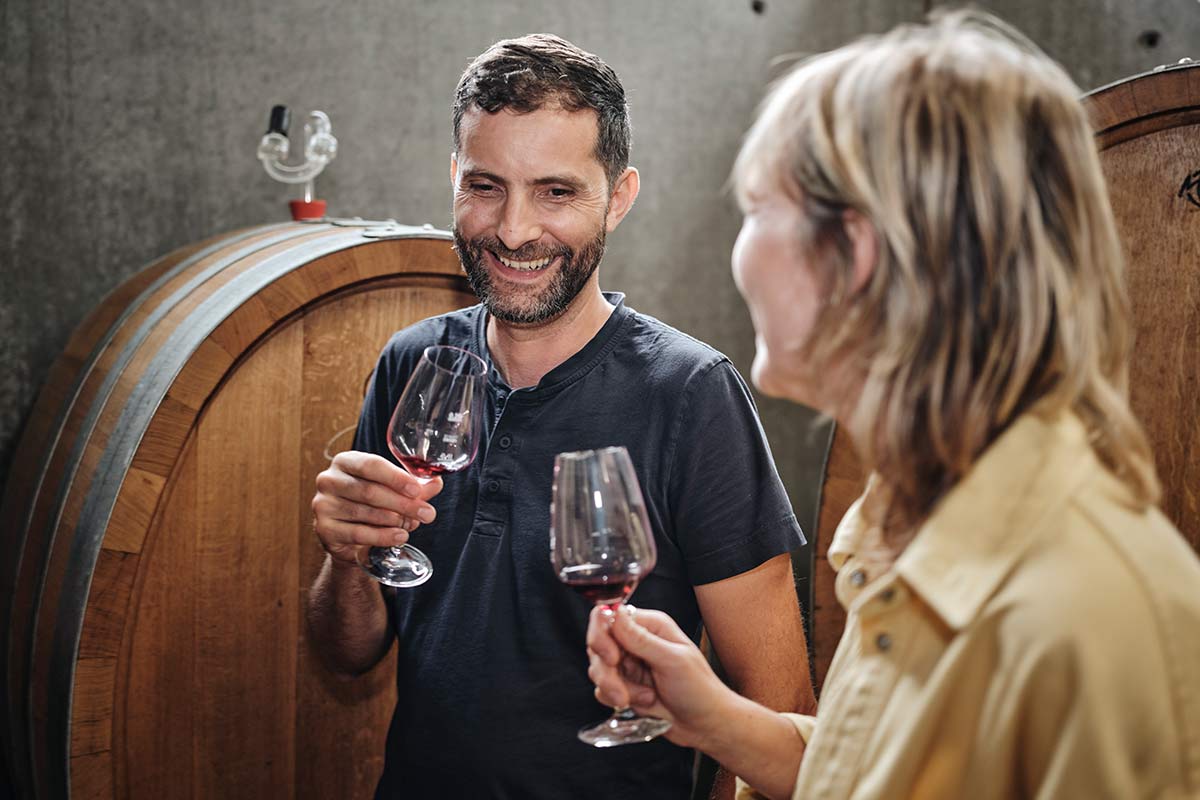 ein Mann und eine Frau halten je ein Glas Wein in der Hand, im HIntergrund sieht man ein Weinfass