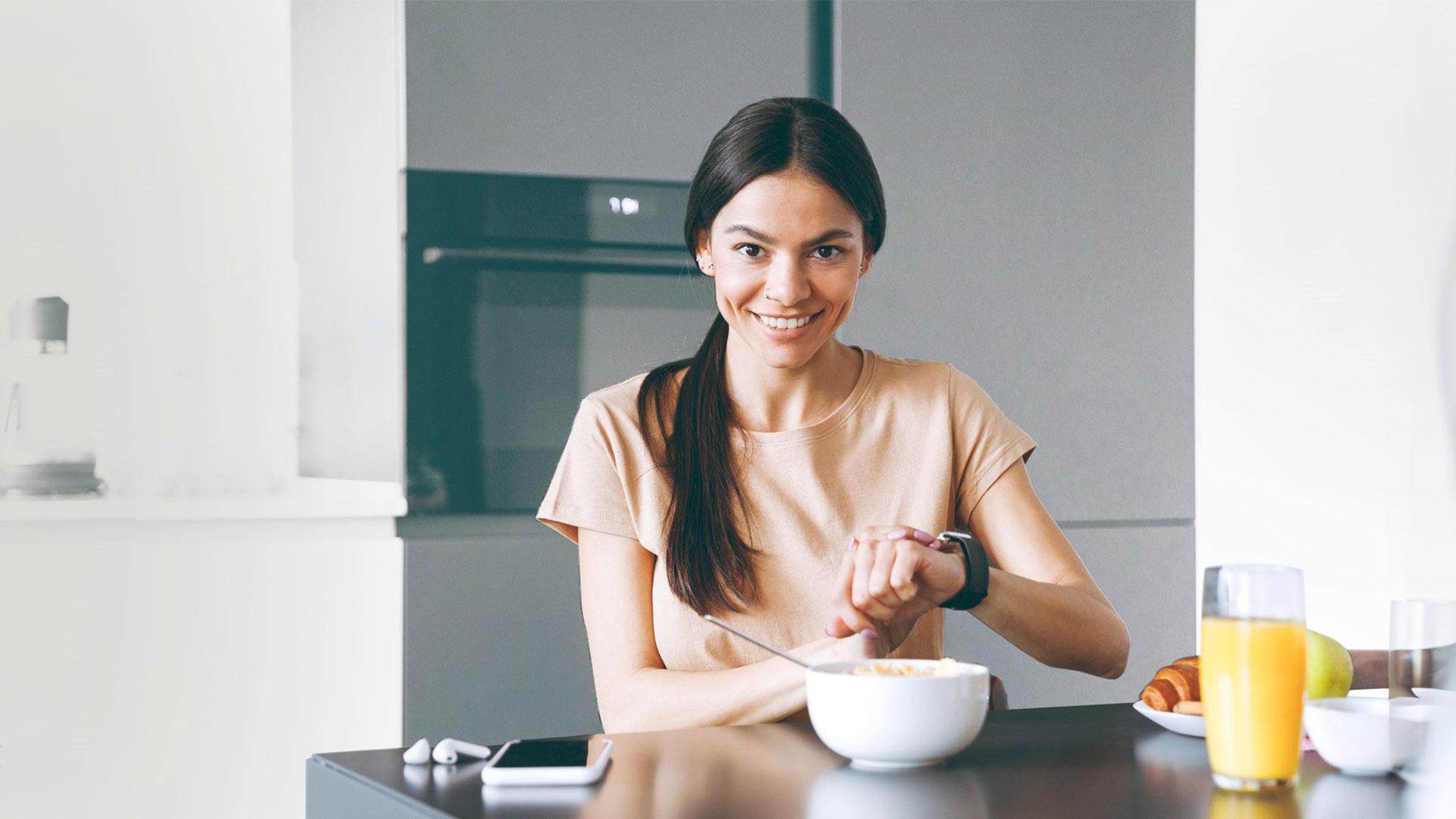 Eine junge Frau sitzt bei einem gesunden Frühstück am Tisch und fasst an ihre Smartwatch