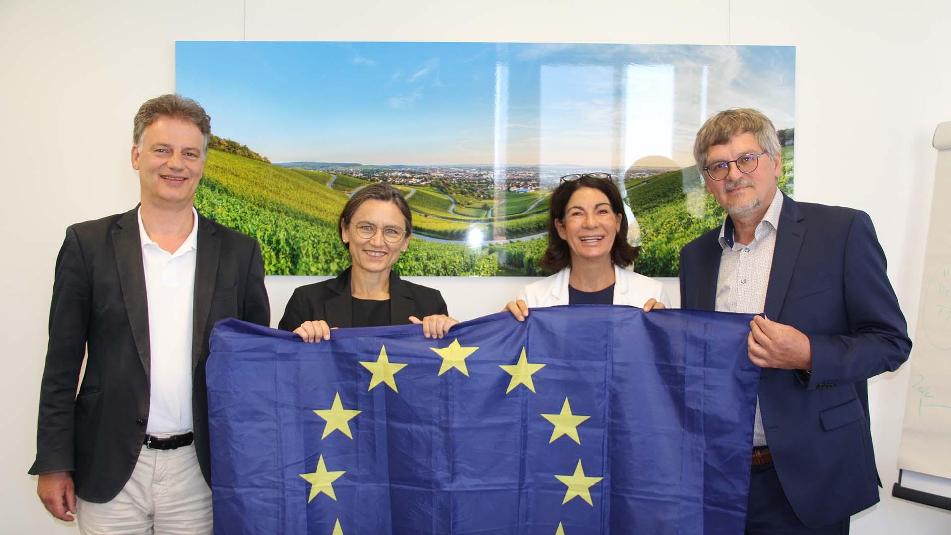 Auf dem Bild stehen vier Personen nebeneinander und halten dabei eine Europaflagge. Von links nach rechts: Prof. Dr. Thomás Bayón, Prof. Dr. Martina Klärle, Prof. Dr. Nicole Graf und Raimund Hudak.