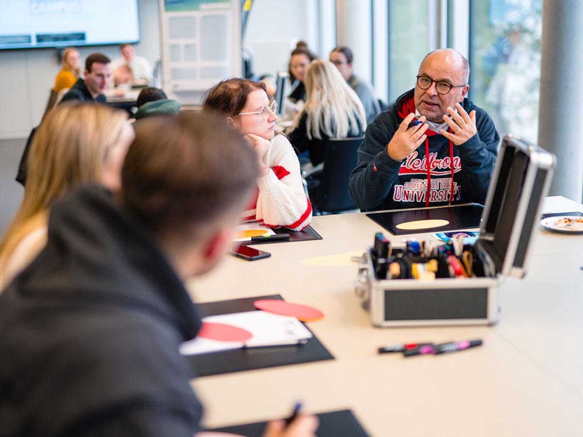 Professor Dr. Stephan Rüschen erklärt Studierenden die Handelskonzepte im neuen Urban Innovation Hub. Alle sitzen am Tisch und diskutieren. 