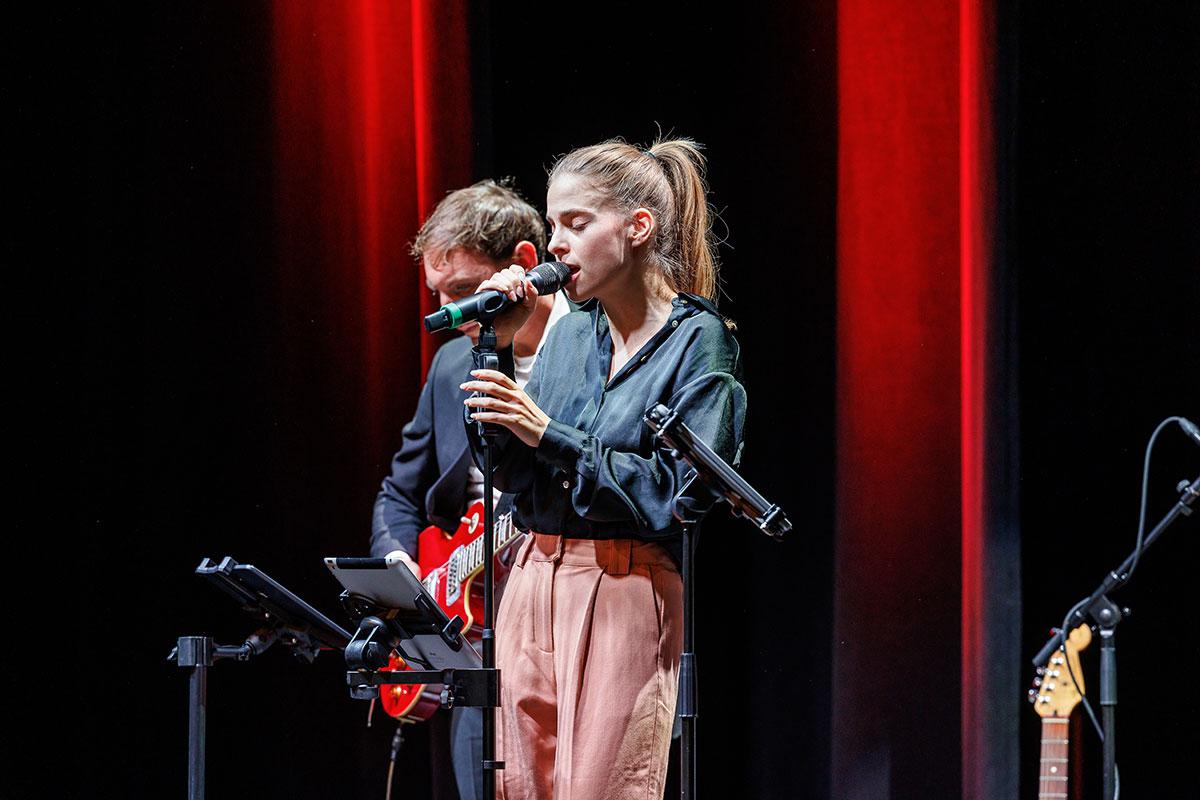 Die Frau Kim Hofmann singt auf der Bühne mit geschlossenen Augen, während ein Mann im Hintergrund E-Gitarre spielt. 