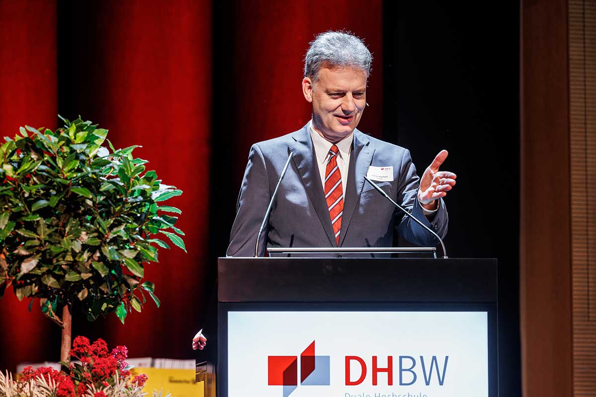 Prorektor Prof. Dr. Tomás Bayòn im grauen Anzug steht am Rednerpult und zeigt mit der Hand zum Publikum. 