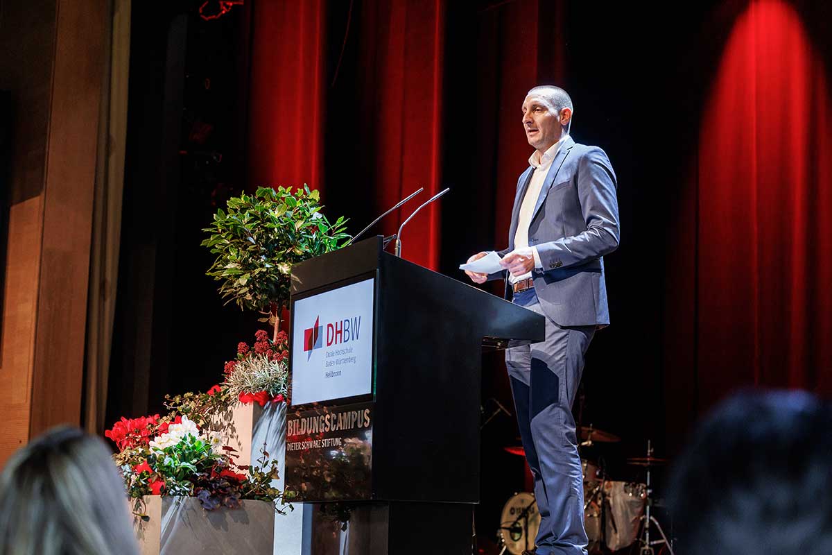 Oliver Vogt steht am Rednerpult, hinter ihm ein rot angeleuchteter Vorhang. 