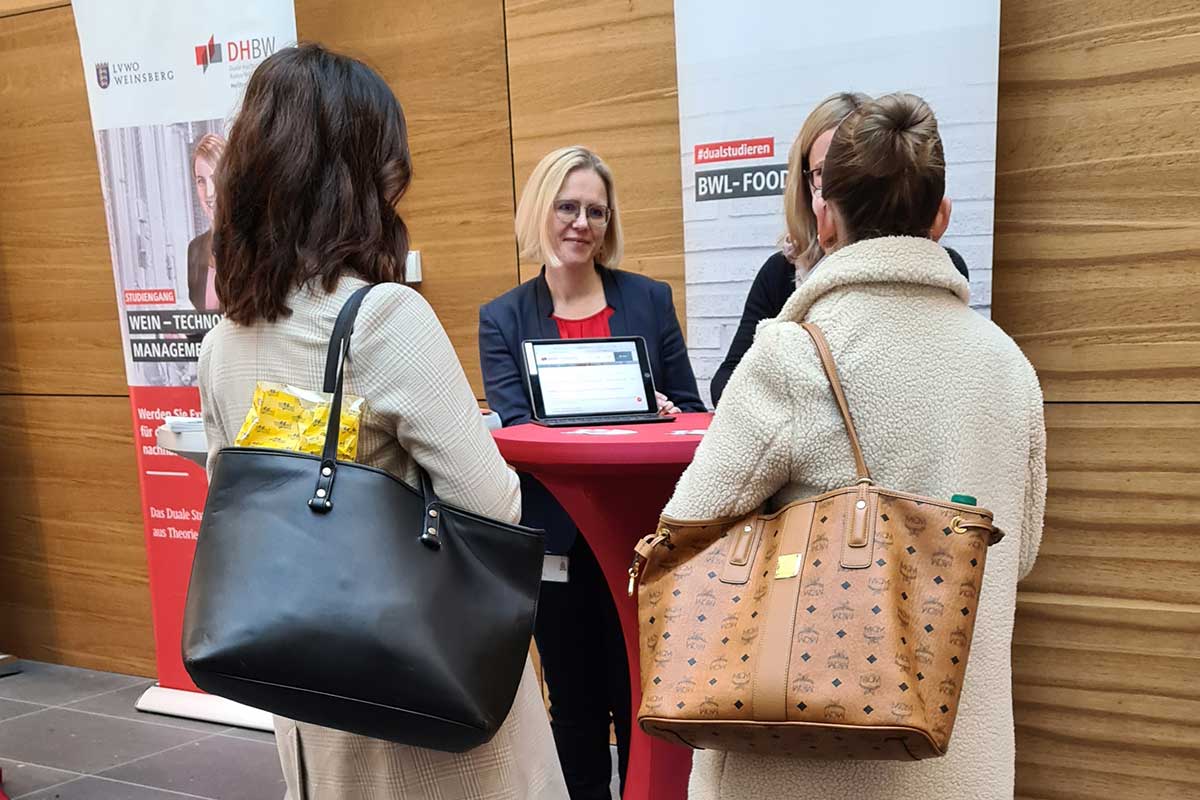 An einem Stehtisch berät eine Mitarbeiterin zwei Schülerinnen zum Studiengang Food Management 