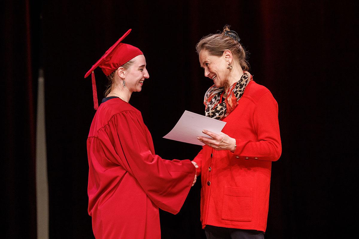 Patricia Wahl in roter Strickjacke überreicht den Preis für Wirtschaft und Ethik an Absolventin in rotem Talar und rotem Barett. 