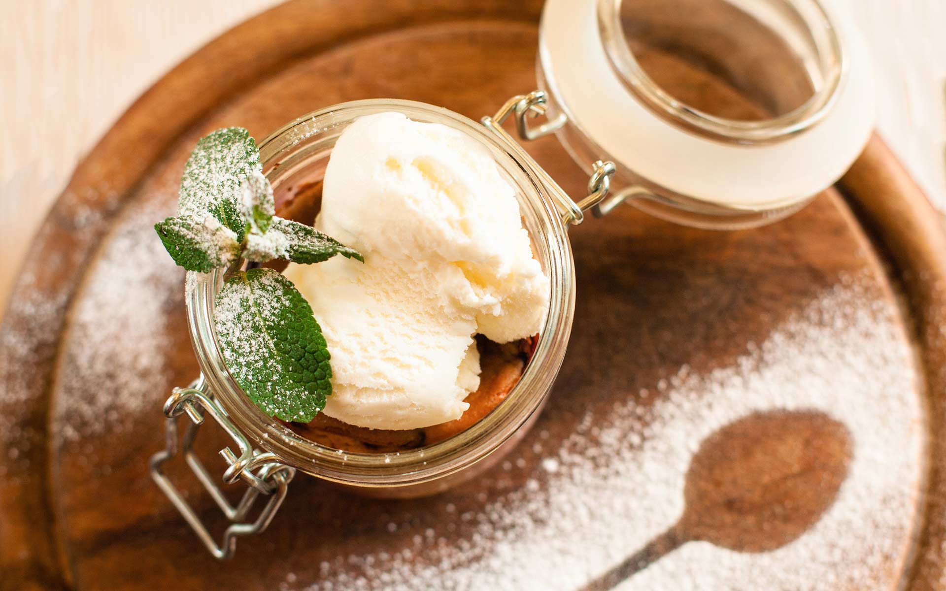 Zwei Kugeln Eis in einem Weckglas mit zwei Minzblättern garniert und mit Puderzucker bestäubt. Neben dem Weck-Glas ist der Abdruck eines Löffels zu sehen. 