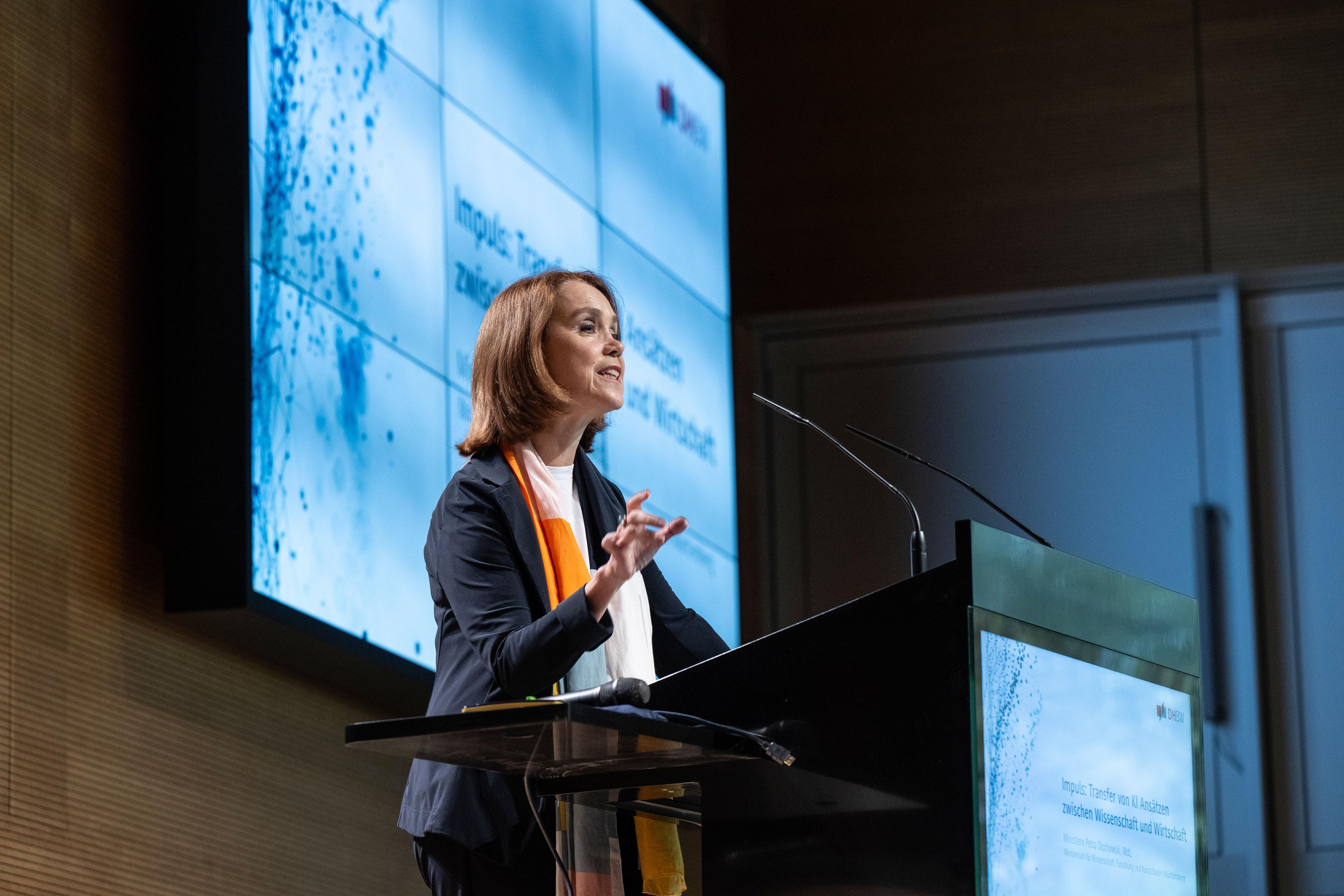 Die baden-württembergische Ministerin für Ministerin für Wissenschaft, Forschung und Kunst Petra Olschowski, MdL, bei der Begrüßung. Sie steht an einem Rednerpult, im Hintergrund eine digitale Leinwand. Olschowski trägt eine dunkelblauen Blazer und ein orange-weiß farbenes Tuch. Beim Sprechen gestikuliert sie lebhaft mit den Händen. 