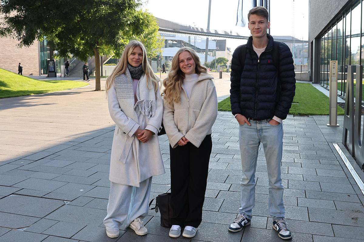 Herzlich willkommen an unsere Erstsemester aus den Studiengängen Handel und Dienstleistungsmanagement. Auf dem Foto sieht man zwei langhaarige junge Frauen und einen jungen Mann mit kurzen Haaren., die neben der Aula stehen. 