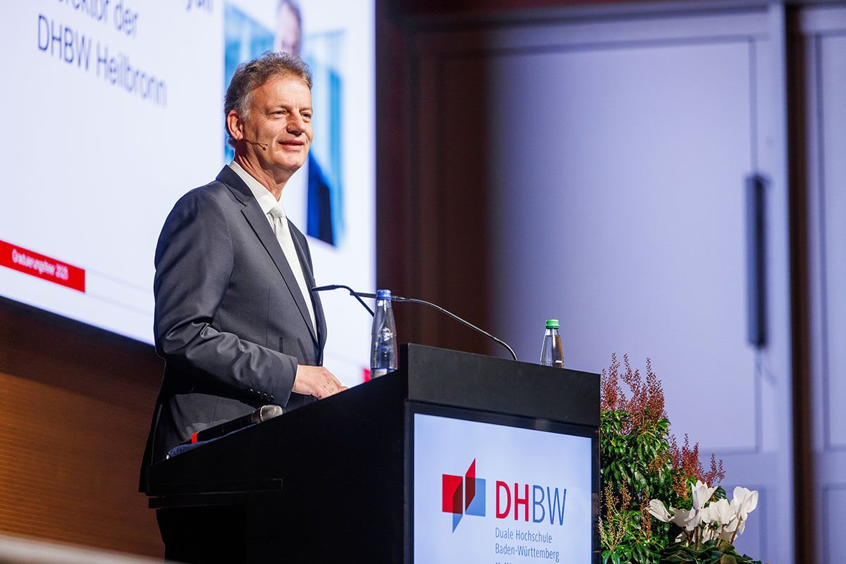 Prorektor Prof. Dr. Tomás Bayón moderierte das Programm der drei Graduierungsfeiern. Prof. Dr. Bayón trägt einen grauen Anzug mit weiißgrauer Krawatte und steht hinter dem Rednerpult. Im Hintergrund sieht man eine Leinwand mit seinem Foto und Namen. 