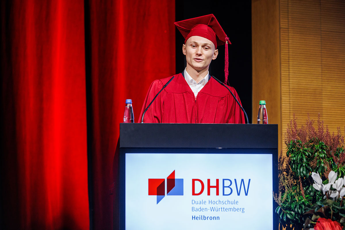 Die drei Absolventen Maximilian Beck (hier im Bild), der ehemalige Sprecher der Studierendenvertretung Klemens Benedikt und Preisträger Felix Kirchgessner ließen gemeinsam mit ihren Kommilitonen die letzten drei Jahre Revue passieren. Maximilian trägt ein rotes Barett und einen roten Talar und steht vor dem roten Vorhang hinter dem Rednerpult auf der Bühne. 