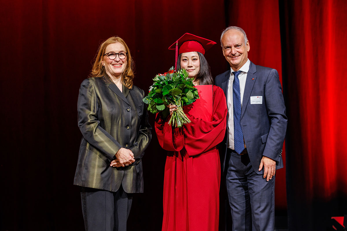 Der diesjährige DAAD-Preis ging an die Südkoreanerin Boram Park, die in Frankfurt ihre Wahlheimat gefunden hat. Im Bild: Leiterin des International Office Verena Kruppa und Prof. Dr. Carsten Kortum. Verena Kruppa hat lange rote Haare, trägt eine moosgrün schimmernde Jacke, Boram Park in rotem Talar und Barett und Prof. Dr. Carsten Kortum mit blauem Anzug. Boram Park hält einen Blumenstrauß in die Kamera.  