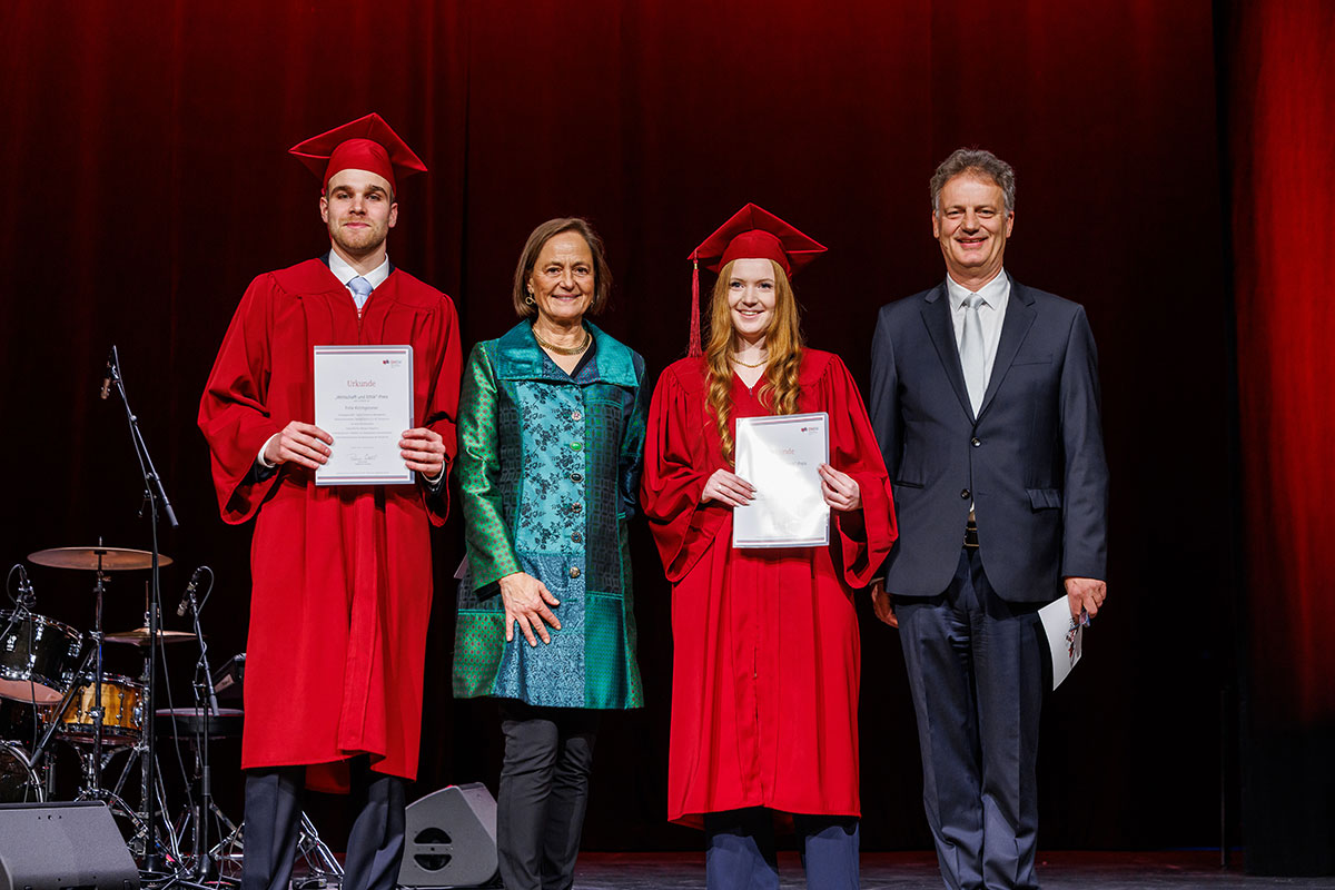 Der diesjährige Preis für Wirtschaft und Ethik wurde an fünf Absolvent*innen für ihre herausragenden Bachelorthesen im Bereich Nachhaltiges Wirtschaften vergeben. Patricia Wahl von der Stiftung für Wirtschaft und Ethik überreichte hier den Preis an Felix Kirchgessner und Jacqueline Rücker. Die beiden Studentinnen tragen rote Talare und Barette, Patricia Wahl eine flaschengrün glänzend schimmernde gemusterte Jacke, Herr Bayón einen grauen Anzug. Die Alumni zeigen ihre Urkunden in die Kamera. 