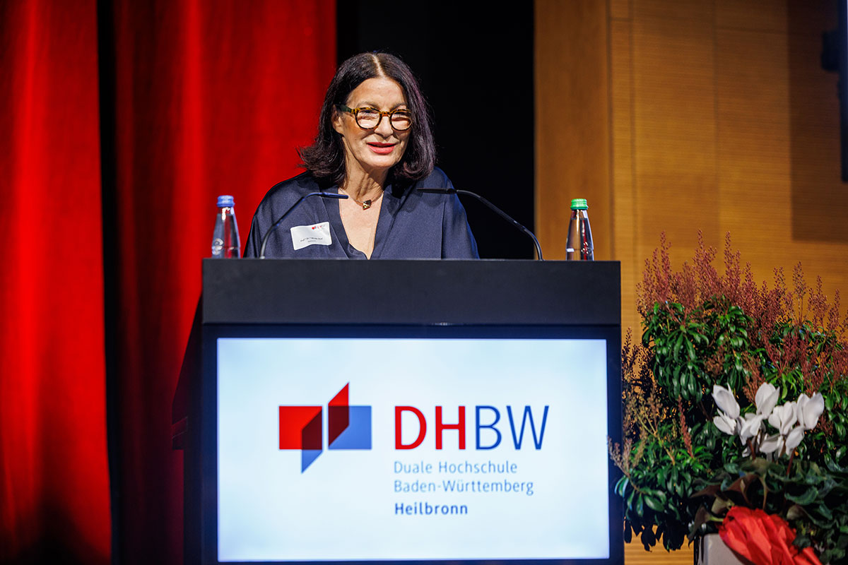 Rektorin Prof. Dr. Nicole Graf begrüßte die Gäste der Graduierungsfeier in der Aula am Bildungscampus. Rektorin Prof. Dr. Nicole Graf steht in einem blauen Anzug hinter einem Rednerpult, auf dem das Logo der DHBW Heilbronn abgebildet ist. Daneben steht ein Blumengefäß auf der Bühne. 