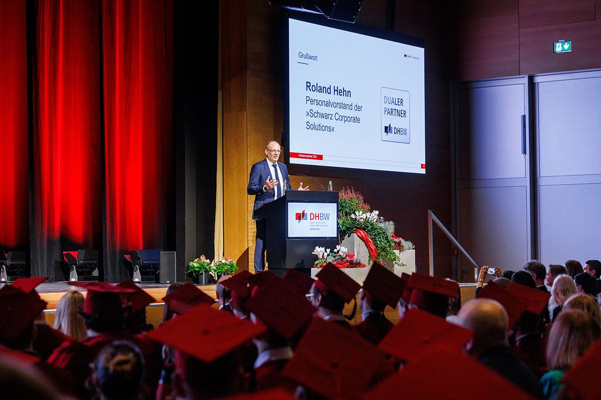 Die zweite Festrede richtete Roland Hehn, Vorstand Personal der SPG Gruppe, an die Alumni aus den Studiengängen BWL-Handel und BWL-Food Management. Herr Hehn steht hinter einem Rednerpult neben der Leinwand, im Vordergrund blickt man über die Köpfe des Publikums in der vollen Aula hinweg. 