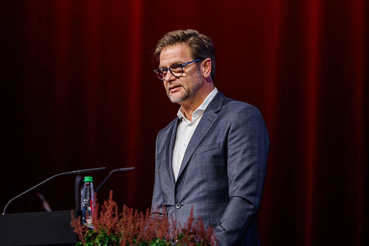 Thomas Storck, CFO der Intersport e.G., gratulierte den Absolvent*innen der Studiengänge BWL-Dienstleistungsmanagement, BWL-Food Management und Wein - Technologie - Management zu ihrem Bachelorabschluss. Thomas Storck trägt einen graublauen Anzug mit weißem Hemd ohne Krawatte, Brille und Dreitagebart. und steht hinter dem Rednerpult. Im Hintergrund sieht man den roten Bühnenvorhang. 