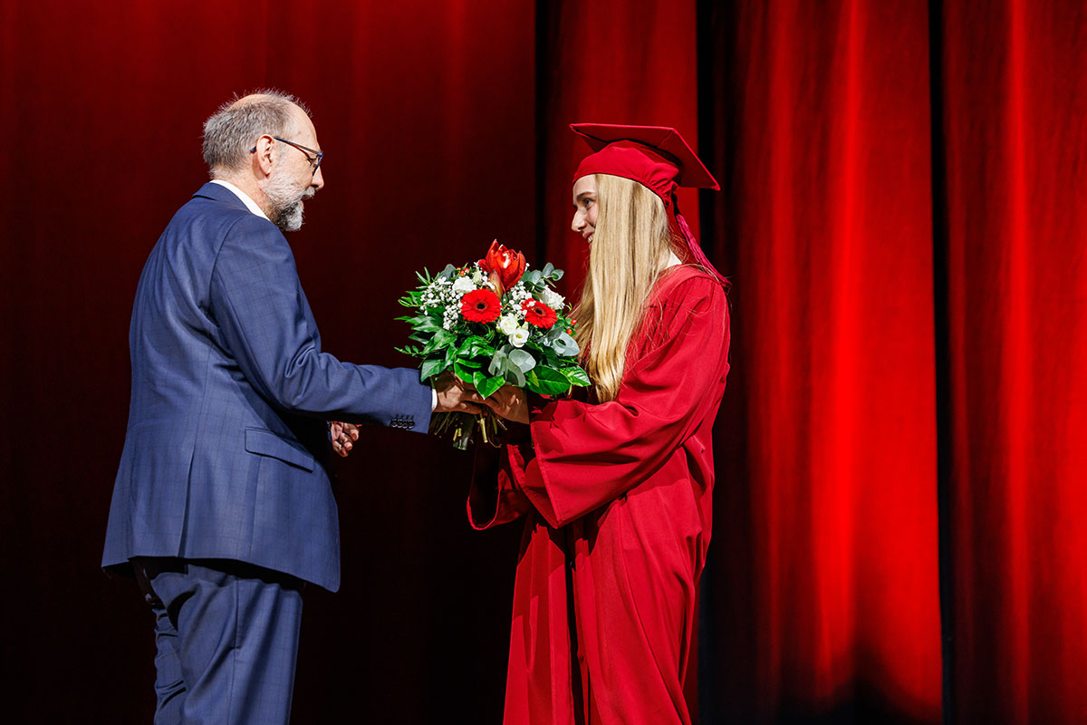 Prorektor Prof. Dr. Tomás Bayón moderierte das Programm der drei Graduierungsfeiern. Wolfgang Jung trägt einen blauen Anzug und überreicht Jasmin - mit langen blonden Haaren in einem roten Talar mit Barrett - einen Blumenstrauß. 