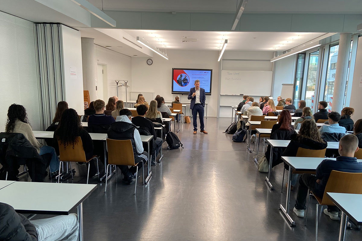 Im Studiengang BWL-Dienstleistungsmanagement können die Studieninteressierten zwischen vier Studienangeboten wählen: Sportmanagement, Media, Vertrieb und Kommunikation, Consulting and Sales und Human Resources Management. Prof. Dr. Niels Biethahn in Anzug und Pullover stellt einem vollen Raum mit jungen Menschen das Studienangebot vor. 