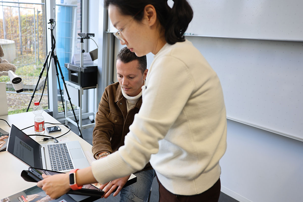 Studieninteressierte testen die digitalen Geräte des Studiengangs Personalisierte Ernährung. Hier im Bild der Biozoom: Durch Handauflegen misst das Gerät die Anzahl der Antioxidantien. Ein junger Mann in brauner Wildlederjacke legt seine Hand auf die Messfläche, die junge Frau Dr. Chenhui Wang stellt das Tablet für die Messung ein. 