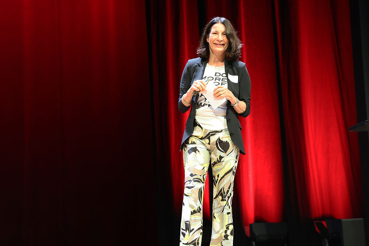 Prof. Dr. Nicole Graf begrüßt die Studierenden. Sie steht auf einer Bühne mit rot angestrahltem Vorhang, trägt eine bunte Hose und ein schwarzes Jacket. 
