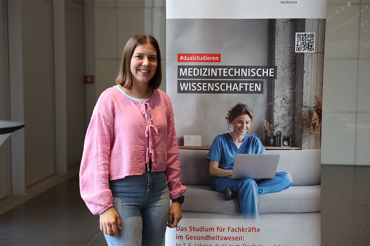 Das Bild zeigt die Studentin Alina Pfenninger vor dem Roll-up des Studiengangs Medinzintechnische Wissenschaften. Sie trägt ein rosa T-Shirt und Jeans.