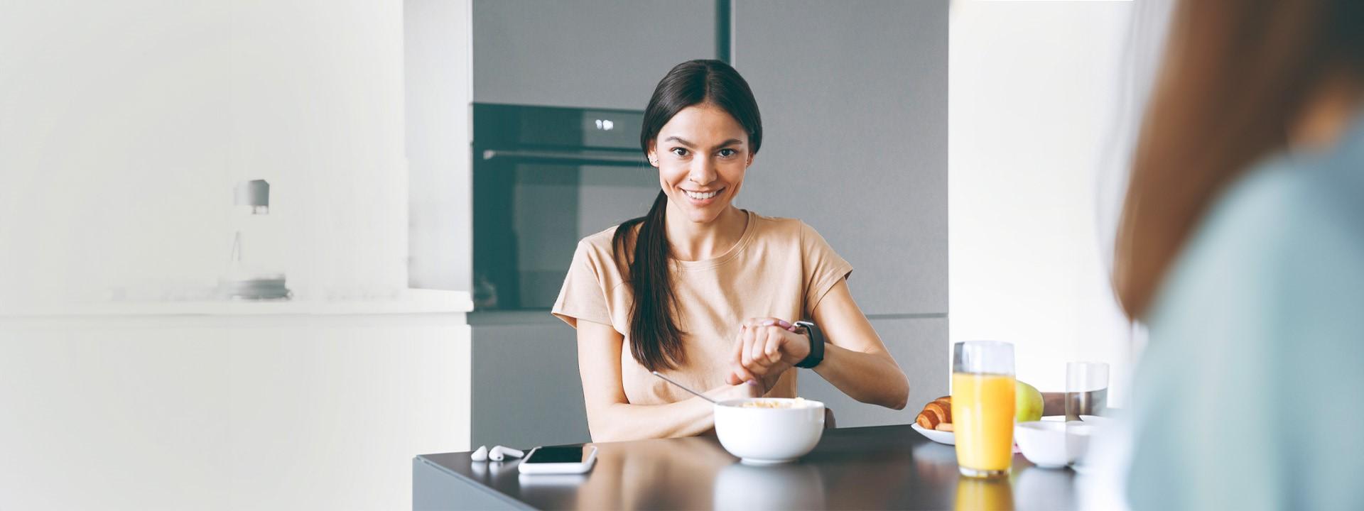 PersE Key Visual Slider web Eine junge Frau sitzt am Tisch mit einem gesunden Müsli vor sich und checkt Daten auf ihrer SmartWatch