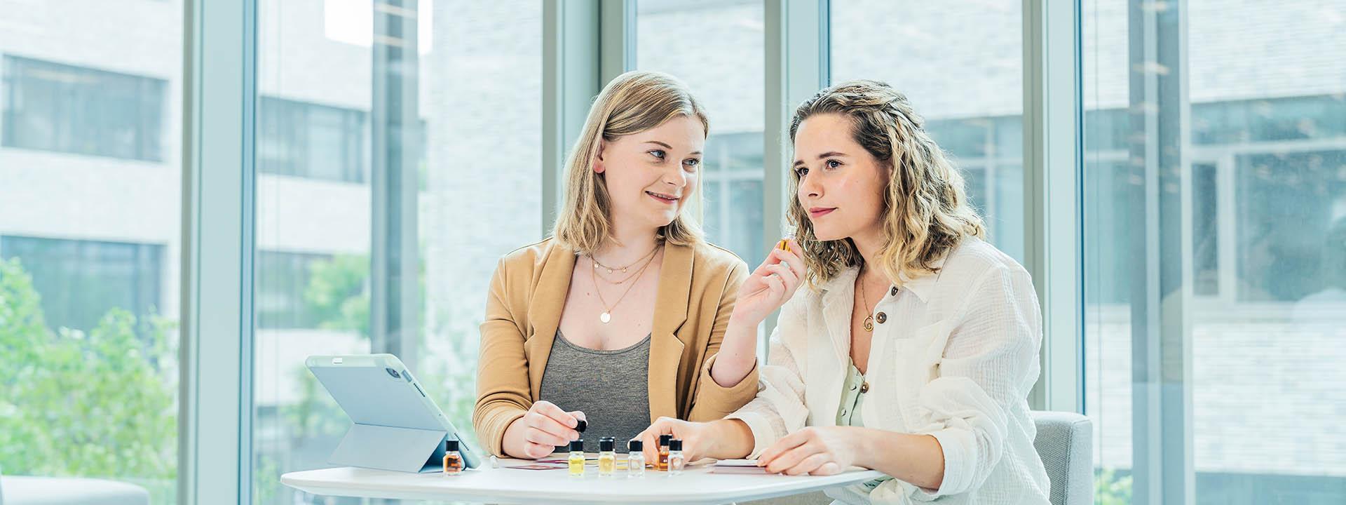 Das Foto zeigt zwei Frauen die an einem Tisch sitzen und kleine Glasfläschchen in der Hand halten. Auf dem Tisch stehen noch sechs weitere Glasfläschchen. Es sieht so aus, als würden sie einen Geruchstest durchführen.
